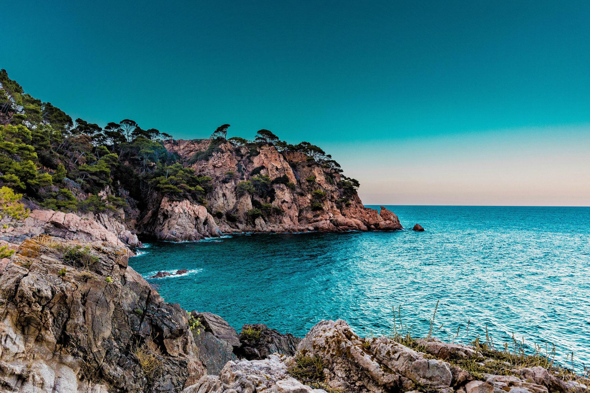Mittelmeer-Küste mit türkisblauem Wasser, steilen Klippen und Pinienbäumen unter klarem Himmel.